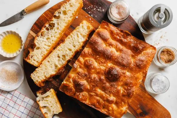 Goldbraune Focaccia mit grobem Salz, bereit zum Belegen. Daneben Olivenöl, Gewürze und ein Messer.