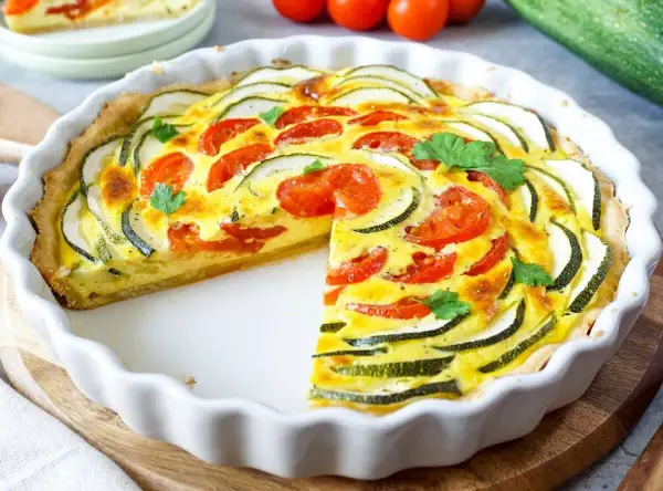 Herzhafte Quiche mit Zucchini und Tomaten, garniert mit Petersilie, in einer weißen Quicheform. Ein Stück fehlt.