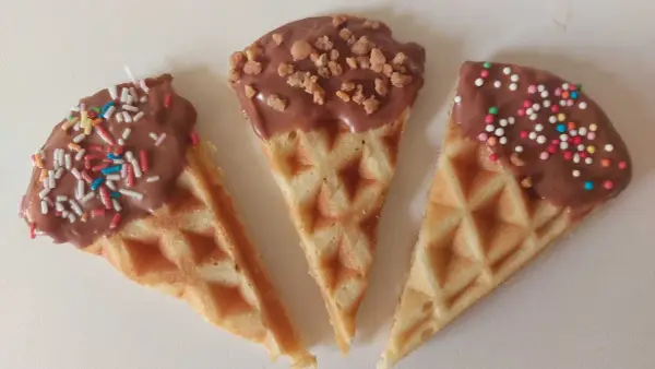 Drei dreieckige Waffeln, mit Schokolade überzogen und bunt bestreut, warten darauf, von Kindern beim Waffeln dekorieren vernascht zu werden.